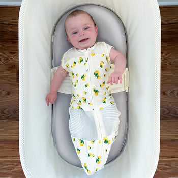 Happy baby in SNOO lying on a gray silk sheet wearing a lemon sleep sack.
