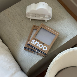 Happiest Baby x Lunya's SNOO Silk Sheets stacked on a side table with SNOObie next to SNOO bassinet.