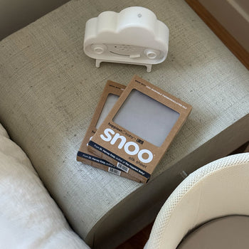 Happiest Baby x Lunya's SNOO Silk Sheets stacked on a side table with SNOObie next to SNOO bassinet.